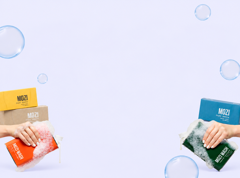 Two hands holding MOZI hand wash bars, one with soapy foam and one dry, with MOZI boxes on either side against a light lavender background with floating bubbles.