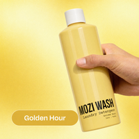 A hand holding a bottle of Mozi Wash Golden Hour Laundry Detergent against a yellow gradient background, with a 'Golden Hour' label overlay.