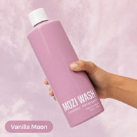 A hand holding a large lavender-pink bottle of MOZI WASH Laundry Detergent in Vanilla Moon scent, set against a matching textured pink background with a color label overlay.