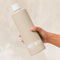 A hand holding a large beige bottle of MOZI WASH laundry detergent against a textured beige background, with a label at the bottom reading 'Signature Cozy'.